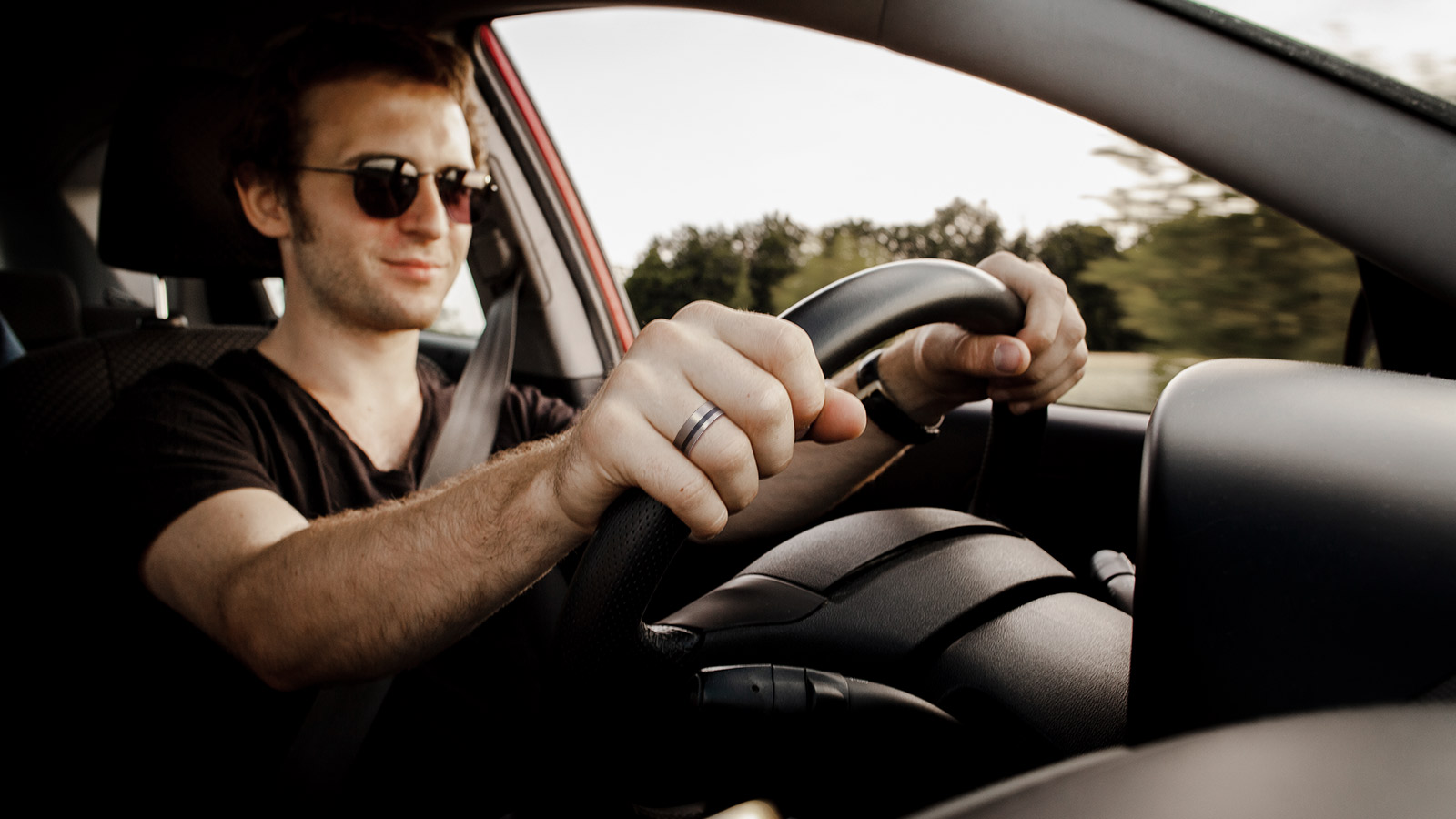 A man in dark glasses is driving a car with a titanium wedding ring on his hand
