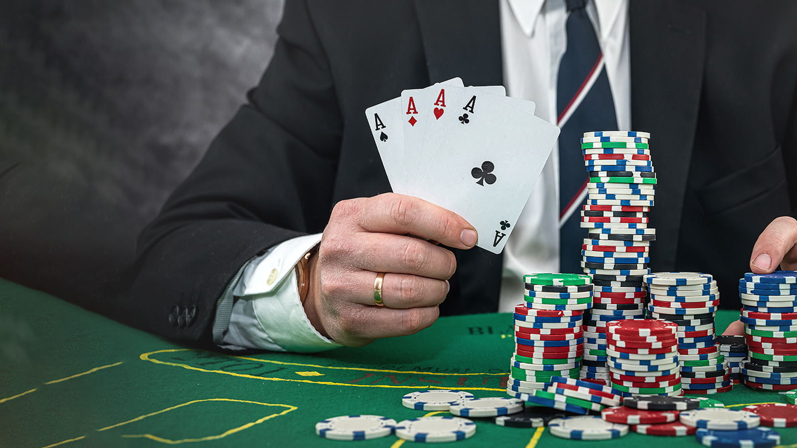 The man in the suit sits at the table with a thick gold ring on his hand, holding a 4 aces playing card and a stack of chips in front of him