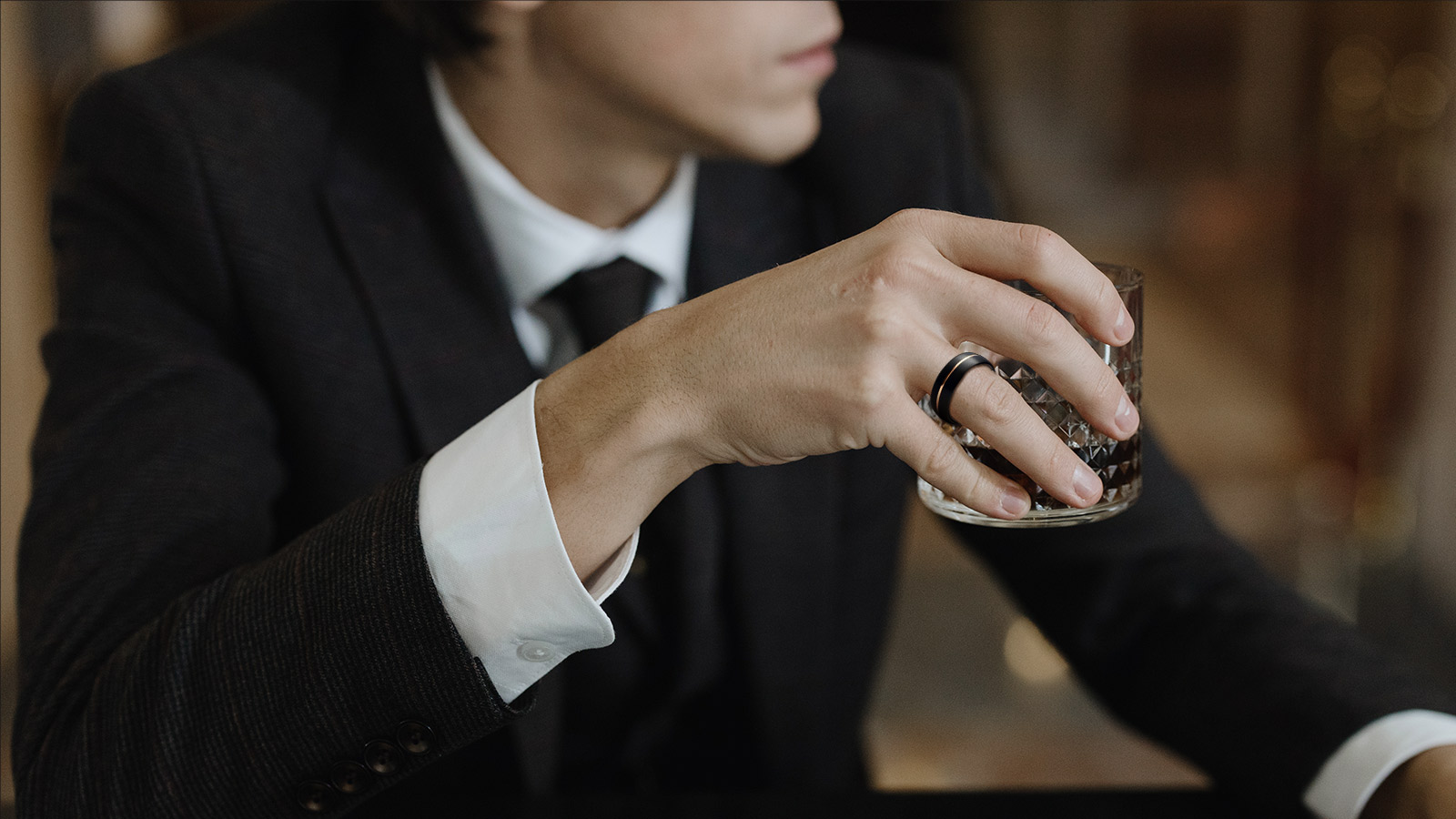 A man in a black suit holds a wine glass and wears a black ring with a gold inlay in the middle