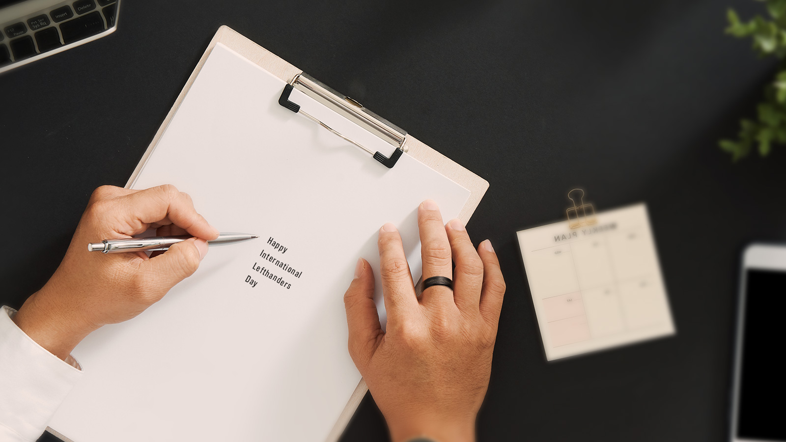 The picture shows a man wearing a thin ring on his right hand, writing on white paper