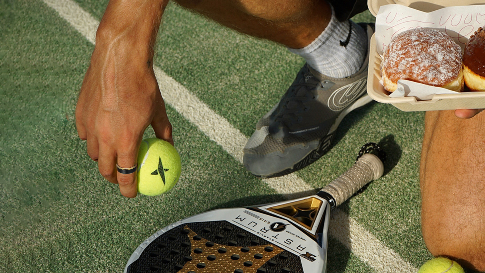 The man is standing on a tennis court, wearing a ring and holding a tennis ball in his right hand and a loaf of bread in his left